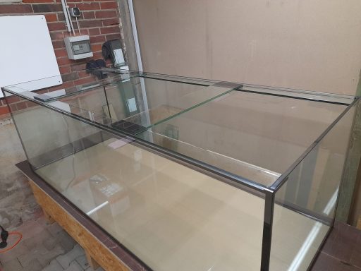 Ein großes, leeres Aquarium mit einem weißen Untergestell steht in einem hellen Raum.